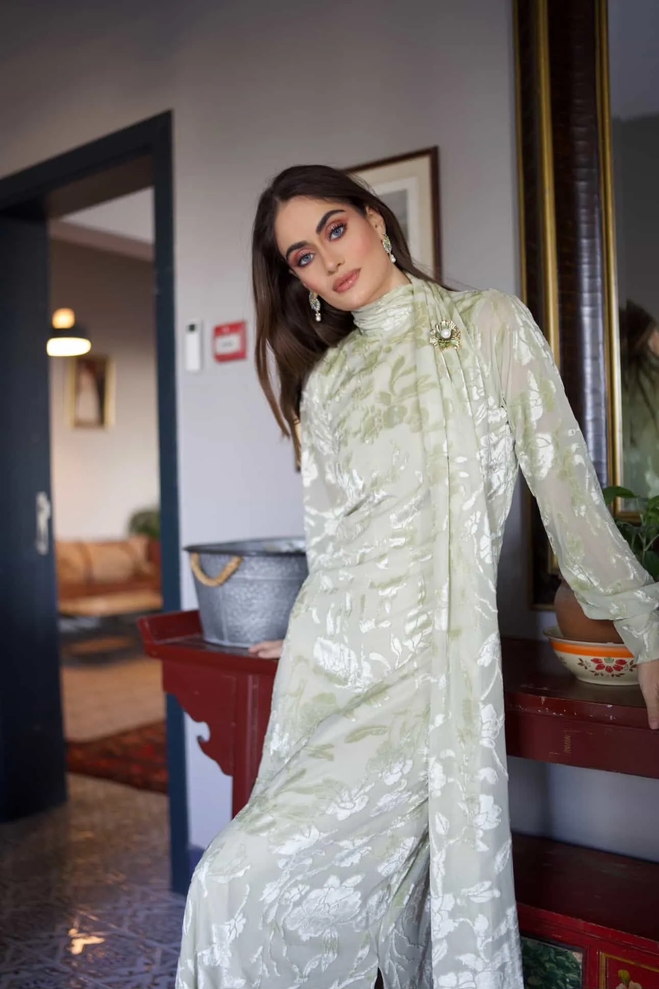 Model in Pastello Mint floral burnout maxi dress with shoulder sash and side slit, leaning against a vintage table.